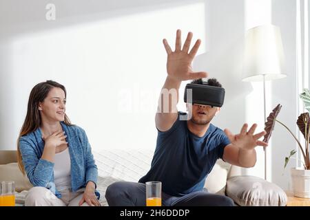 Young man enjoying playing video game in virtual reality glasses at home, his girlfriend sitting near by Stock Photo