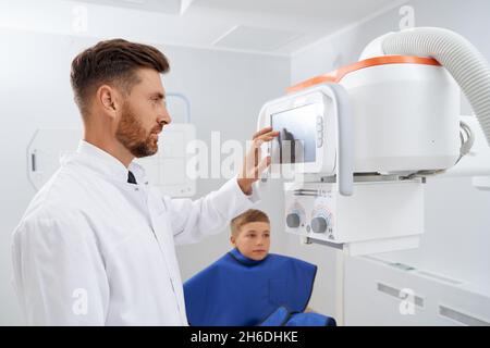 Side view of brunette doctor with beard touching screen of usg modern tool, starting procedure of diagnostic. Professional radiologist doing ultrasound diagnostic for little boy wearing blue robe.  Stock Photo
