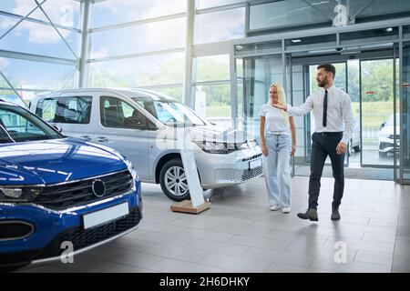 Caucasian handsome man and beautiful woman walking together at modern auto salon. Salesman helping female customer buying new transport. Stock Photo