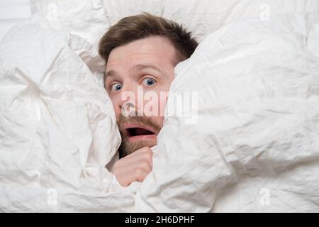 Caucasian man woke up from a nightmare, he looks out from under the blanket in fright and looks with big eyes. Top view. Stock Photo