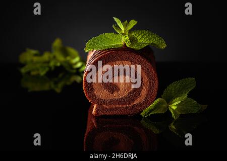 Chocolate roll cake with mint on a black reflective background. Stock Photo