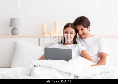 Happy european millennial man and woman hugging, sitting on bed, use laptop in white bedroom interior Stock Photo