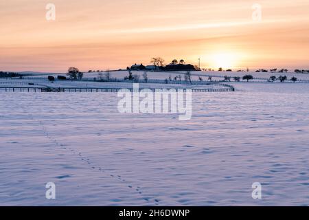 Farm buildings and wind turbine on skyline behind Hexham racecourse all covered with snow and silhouetted against a vivid orange sky at sunset. Stock Photo