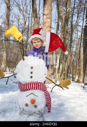 child boy in Santa hat and homemade funny snowman standing upside down on his head. Christmas festive season in a snowy forest. Winter walks and fun w Stock Photo