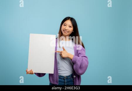 Place for text. Cheerful asian lady pointing at empty banner with free space for your design, blue background, mockup Stock Photo