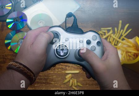 Close-up of hands holding a gray joystick from the console against the background of disks and fries scattered on the floor Stock Photo
