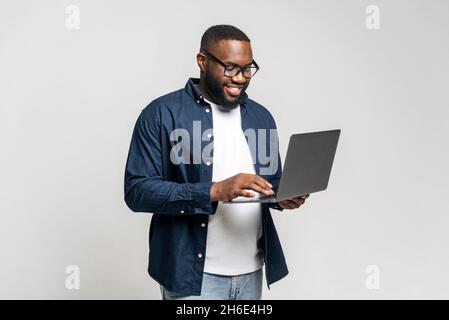 Portrait of young African American guy standing isolated on grey wall holding laptop, typing messages, chatting with friend on social media online, watching video, enjoying new application, websurfing Stock Photo