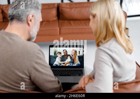 Middle-aged couple using laptop for video connection with grown children and grandchildrens, grey-haired man and blonde woman talks with multiracial family on the screen. Video chatting concept Stock Photo