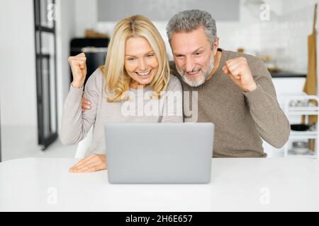 Overjoyed middle-age couple win in online lottery, received good news, happy mature husband and wife staring at the laptop screen and screen yes happily, raising hand in triumph and victory Stock Photo