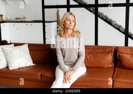 Portrait of serene and carefree beautiful middle-aged woman sitting on the couch and looks away lost in thoughts and dreams. Peaceful well-looking lady resting at home alone Stock Photo