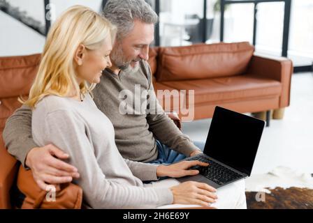 Advertising, mock-up concept. Middle-aged couple sitting in embraces on the sofa at home and using laptop with a blank empty display, grey-haired man and blonde woman looking at screen. Template Stock Photo