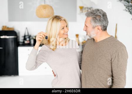 Moving day concept. Happy middle-aged couple in love holds keys from new house, looking to each other happily, glad with achievement, senior owners of real estate Stock Photo