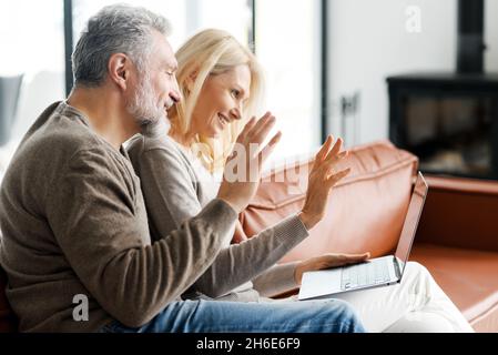 Positive middle-aged couple is using laptop for video call sitting on comfortable sofa in cozy living room. Cheerful spouses waving into webcam. Online communication concept Stock Photo
