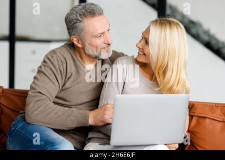 Middle-aged couple sitting on the couch and using laptop for spending leisure together, watching movies, web surfing, shopping online. Cheerful senior spouses with a laptop at home look at each other Stock Photo