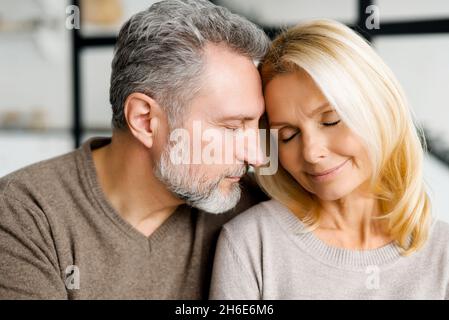 Gentle portrait of romantic serene middle-aged couple in embraces at home. Mature spouses in love sitting on the sofa with eyes closed, feels appeasement and calm. Bonding and love concept Stock Photo