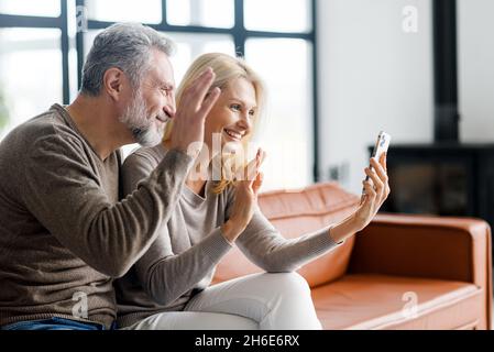 Cheerful middle-aged couple using smartphone for video connection with family or friends on distance, happy mature man and mid-age woman holding mobile phone and waving, making video call, side view Stock Photo