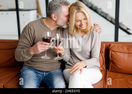 Happy marriage anniversary. Cheerful middle-aged spouses celebrating anniversary at home, sitting on the sofa and holding glases with wine, enjoying time together, laughing Stock Photo