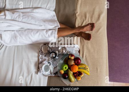 Crop from above view of legs of woman in white morning gown lying on bed with plate full of fresh fruits and coffee set placed near Stock Photo