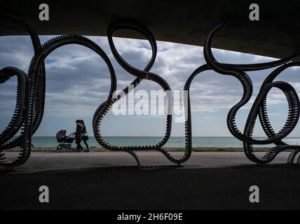 Members of the public walk past the The Long Bench at Littlehampton.The Long Bench is thought to be the longest bench in Britain and one of the longest in the world. The wood and stainless steel bench â€˜flowsâ€™ along the promenade at Littlehampton in West Sussex - curving round lamp posts and obstacles, twisting up into the seafront shelters, dropping down to paths and crossings. Stock Photo