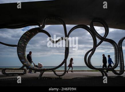 Members of the public walk past the The Long Bench at Littlehampton.The Long Bench is thought to be the longest bench in Britain and one of the longest in the world. The wood and stainless steel bench â€˜flowsâ€™ along the promenade at Littlehampton in West Sussex - curving round lamp posts and obstacles, twisting up into the seafront shelters, dropping down to paths and crossings. Stock Photo