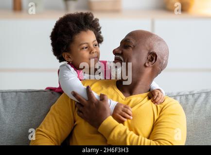 Cheerful little black girl play with older grandfather from back, hugging man at home interior Stock Photo