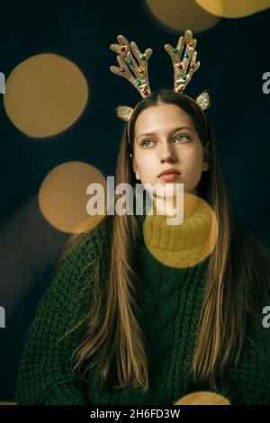 A beautiful teenage girl in Christmas antlers and a green sweater dreamily looks up to the side. Close-up, vertical. Dark background with bokeh from g Stock Photo