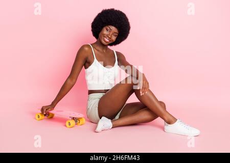 Full size photo of lovely millennial curly hairdo lady sit with skate wear white top shorts isolated on pastel pink color background Stock Photo