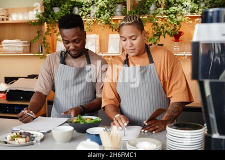 Horizontal medium portrait of young African American man and woman working in small cafe kitchen creating new menu making notes Stock Photo