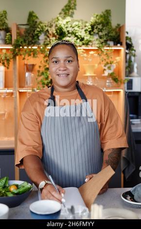 Vertical medium portrait of young adult Black woman working in small cafe kitchen writing down list of products in notebook looking at camera Stock Photo