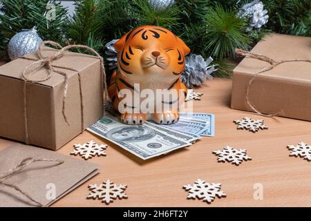 A piggy bank in the shape of a tiger standing on cash dollars on a wooden background with gift boxes, snowflakes and fir branches. Success and wealth Stock Photo