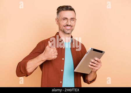 Photo of smart middle-aged guy use app gadget show thumbup symbol great covid suggest isolated over pastel beige color background Stock Photo