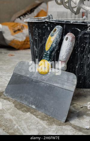Two trowels standing on a concrete floor leaning on a black plastic bucket. Empty paper bags of plaster are lying in the background. Room renovation c Stock Photo