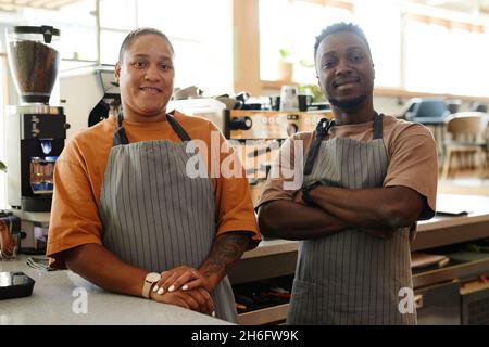 Horizontal medium portrait of modern young co-workers wearing aprons standing at cafe counter against coffee machines looking at camera waiting for cu Stock Photo