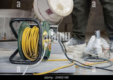 Yellow electric extension cord on a green spool. Four plugs are plugged into the extension cord. In the background are men's feet, buckets and bags of Stock Photo