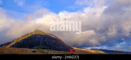 A woman in an orange jacket taking a photograph of a rescue helicopter flying over the Carneddau mountains in Snowdonia National park, North Wales, UK Stock Photo