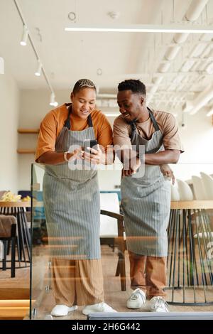Vertical full length shot of young African American man and woman working together in modern cafe watching something funny on smartphone during break Stock Photo