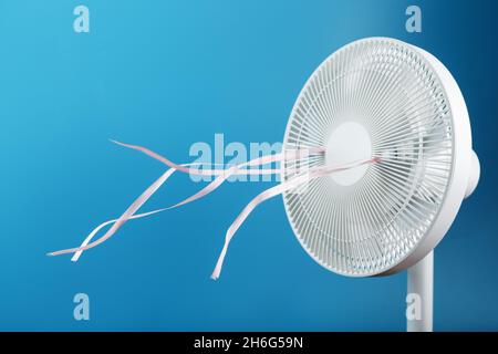 The electric fan is white with pink ribbons fluttering in the wind on a blue background. Free space, minimalistic style. Stock Photo