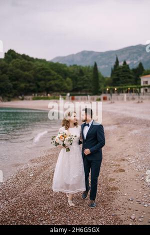 Groom hugs bride in a white dress with a bouquet of flowers, walking with her along the pebble beach against the backdrop of mountains and green trees Stock Photo