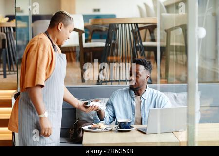 Young Black man having lunch in modern cafe sitting at table paying for order with bank card Stock Photo