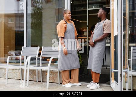 Horizontal full shot of young Black man and woman wearing aprons standing at cafe entrance talking about something Stock Photo