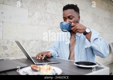Portrait of handsome young African American man wearing casual outfit sitting at cafe table outdoors using laptop and drinking coffee Stock Photo