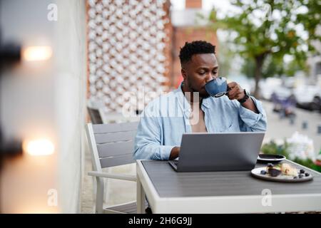 Portrait of young Black man wearing stylish casual clothes sitting at cafe table outdoors working on laptop and drinking cappuccino Stock Photo