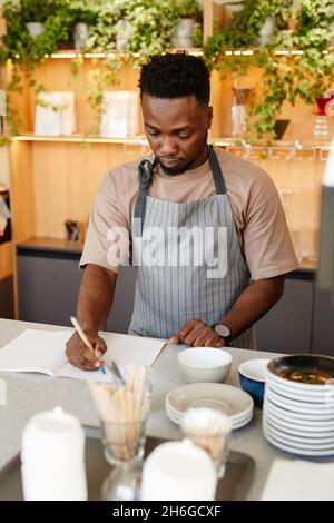 Vertical medium portrait of young African American man wearing apron standing at cafe kitchen table writing something in notebook Stock Photo