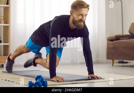Happy athletic young man doing plank with knee tucks during fitness workout at home Stock Photo