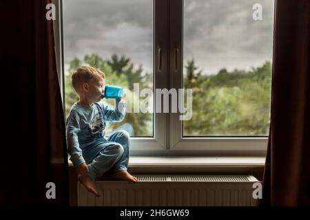 Blonde boy in blue pajamas drinking milk and sitting on windowsill Stock Photo