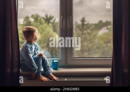 Side view of boy in blue pajamas sitting on windowsill Stock Photo