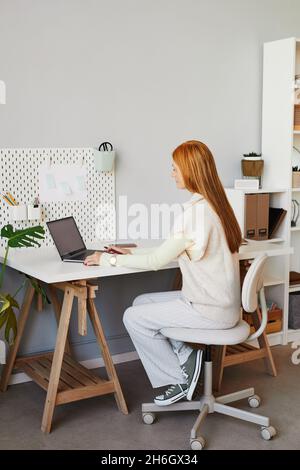 Vertical full length portrait of young red haired woman using laptop or studying at minimal home workplace Stock Photo