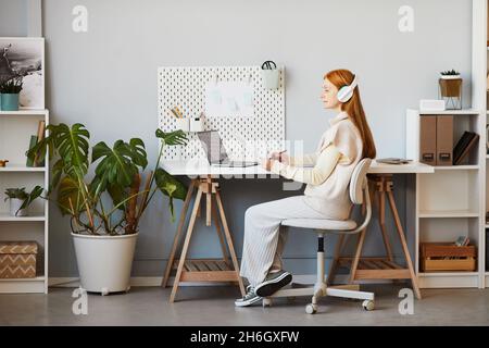 Full length portrait of red haired woman using laptop or studying at minimal home office workplace, copy space Stock Photo