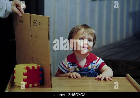 Austin Texas USA, 1995: Child development: 10-month old baby boy shows lack of 'object permanence' during an experiment in a college laboratory, as a lab assistant hides a toy behind a piece of cardboard.  MR EC-0000  ©Bob Daemmrich Stock Photo