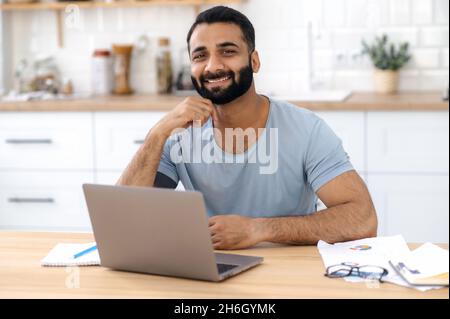 Portrait of charismatic positive young adult Indian man, freelancer, designer or IT specialist, works from home using laptop, looking directly into camera and smiling friendly Stock Photo